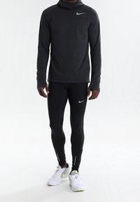 Haut de sport à manches longues gris foncé avec capuche, leggings noirs moulants et baskets blanches avec des accents verts. Marque Nike affichée.