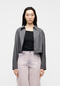 Νεαρή γυναίκα που φορά cropped γκρι μπουφάν πάνω από μαύρο τοπ και ψηλόμεσα ανοιχτόμωβ τζιν, στέκεται μπροστά σε α πλό λευκό φόντο.