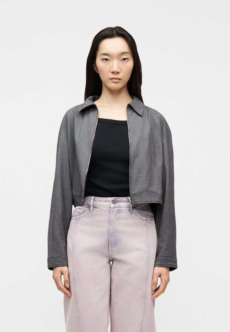 Νεαρή γυναίκα που φορά cropped γκρι μπουφάν πάνω από μαύρο τοπ και ψηλόμεσα ανοιχτόμωβ τζιν, στέκεται μπροστά σε απλό λευκό φόντο.