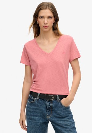 Woman wearing a pink V-neck short-sleeve shirt tucked into blue jeans with a black belt, standing with one hand in pocket.