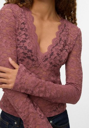 Femme portant un haut à manches longues en dentelle florale mauve avec un col en V festonné, vue des épaules à la taille, bras croisés sur la poitrine.