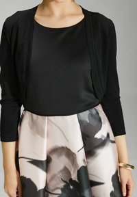 Cardigan negro ajustado con mangas tres cuartos sobre una blusa negra brillante. La falda color rubor presenta patrones florales negros y plisados. Pulsera dorada visible.