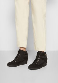 Person som bär off-white, korta byxor och svarta kängor med kilklack, snörning och sidodragkedja, står på ett ljusgrått golv.