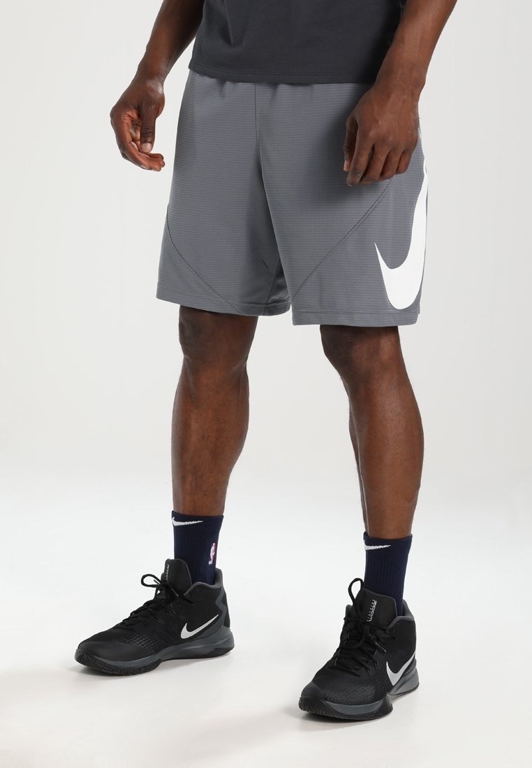 Personne portant un short Nike gris avec un grand logo blanc, des baskets montantes noires Nike et des chaussettes foncées, debout sur un fond blanc uni.
