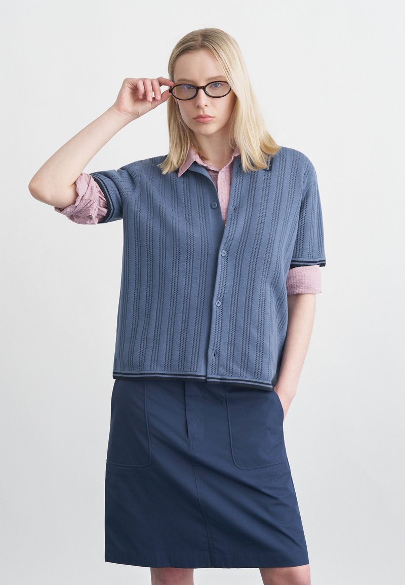 Ξανθιά γυναίκα που προσαρμόζει μαύρα γυαλιά, φορώντας μπλε ζακέτα με κουμπιά και κοντά μανίκια πάνω από ροζ πουκάμισο και σκούρο μπλε φούστα, στέκεται μπροστά σε λευκό φόντο.