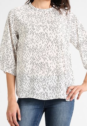 Kobieta ubrana w białą, luźną bluzkę z czarnym abstrakcyjnym wzorem kropek oraz rękawami do połowy przedramienia, w połączeniu z niebieskimi dżinsami.