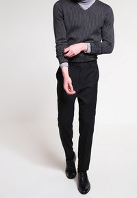 Pantalon noir sur mesure associé à un pull ajusté à col en V sombre à motif. Le modèle porte des chaussures noires, affichant une silhouette épurée et moderne.