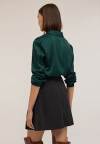 Bluză de satin verde închis cu mâneci răsucite, asociată cu o fustă plisată neagră. Outfit-ul are un guler clasic și o siluetă elegantă.