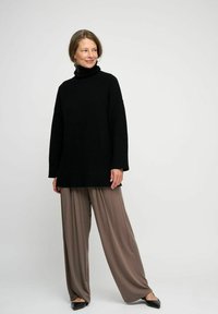 Svart stickad turtleneck-tröja med lös passform och texturerat mönster, kombinerad med vida taupe-byxor och svarta platta skor.