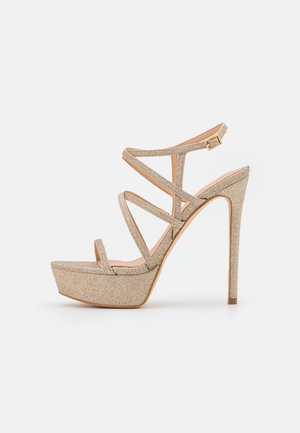 Sandalia de tacón alto con brillo dorado, suela con plataforma, múltiples tiras delgadas y cierre de hebilla en el tobillo, vista lateral sobre fondo blanco.