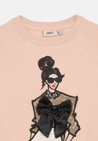 Světle růžová mikina s ilustrací ženy v brýlích, doplněná černou mašlí a texturovanými akcenty.