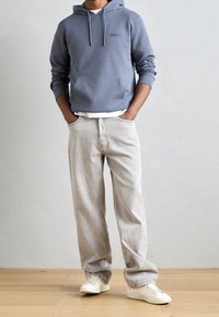 Felpa blu con tasca frontale e cordini, abbinata a jeans larghi grigi chiari e sneakers bianche su un pavimento di legno.