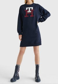 Tmavě modré šaty ve stylu mikiny s velkým texturovaným logem "TOMMY" v červené a bílé barvě. Mají dlouhé rukávy a kulatý výstřih, nošeny s tmavě modrými botami.