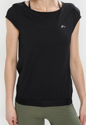 Femme portant un haut de sport noir ample à manches courtes sur des bretelles visibles de soutien-gorge de sport et des leggings moulants verts olive.