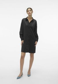 Czarna, oversize koszulowa sukienka z kołnierzem, długimi rękawami i gładką, błyszczącą teksturą. W połączeniu z jasnoniebieskimi, spiczastymi szpilkami.