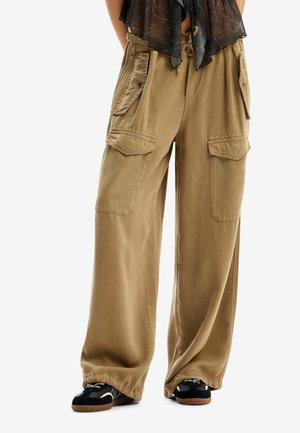 Pantalon cargo ample beige avec grandes poches à rabat et ourlets élastiqués, porté avec des baskets noires et blanches.
