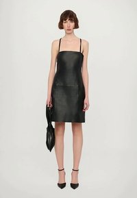 Sieviete, tērpusies melnā rokām bez piedurknēm ādas kleitā ar plānām siksnām, tur melnu ādas somiņu un valkā melnas augstpapēžu kurpes.