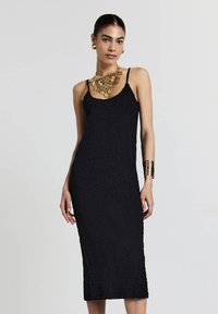 Schwarzes Midikleid mit strukturierter Oberfläche, Spaghetti-Trägern und taillierter Silhouette. Akzentuiert mit einer goldenen Statement-Halskette und einem Armband.