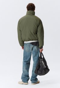 Oliven grøn pufferjakke, baggy blå jeans, beige støvler, holder en sort taske med et ternet tilbehør. Fleecefor er synligt ved taljen.