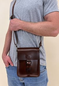 Borsa a tracolla in pelle marrone con chiusura a gancio, tracolla regolabile e dettagli cuciti. Forma rettangolare compatta.