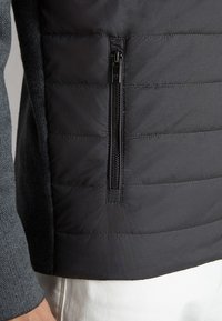 Sezione di giacca trapuntata nera con tasca con zip verticale, indossata su maniche grigie testurizzate e pantaloni bianchi.