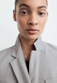 Detailný záber osoby s hnedými očami, ktorá má na sebe svetlosivý blazer a čierny top na jednoduchom svetlom pozadí.