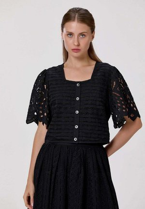 Jeune femme aux cheveux bruns et raides porte un haut en dentelle noire boutonné avec des manches bouffantes et une jupe assortie, debout devant un fond uni.