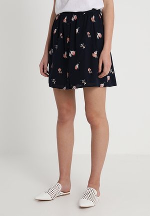 Femme portant une jupe fleurie bleu marine avec des boutons à l'avant, un haut blanc et des chaussures blanches à enfiler perforées, debout sur un fond uni.
