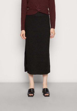 Kobieta ubrana w czarną prążkowaną midi spódnicę z bocznym rozcięciem, bordowy sweter z długim rękawem oraz czarne mule na obcasie z odkrytymi palcami.