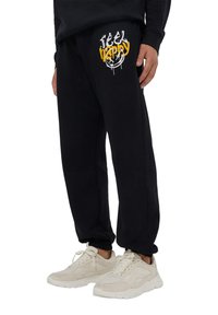 Pantaloni sportivi neri con grafica "Feel Happy" in bianco e giallo, dotati di una vita elastica e polsini. Accoppiati con sneaker beige.