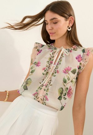 Frau mit langen braunen Haaren trägt eine ärmellose Bluse mit Blumenmuster und Rüschen sowie einen weißen Rock, kombiniert mit einem goldenen Armband und Ohrringen.