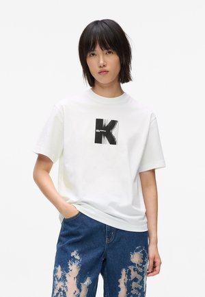 Valkoinen puuvillapaita, jossa on musta graafinen "K"-kuvio rintamalla. Mallilla on päällään kuluneet siniset farkut, joissa on näkyvissä paikkaus.