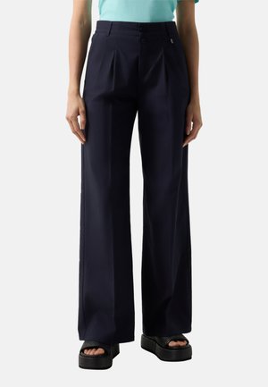 Frau trägt hochtaillierte, marineblaue, weit geschnittene Hosen mit Vorderfalten und schwarzen Plateausandalen, steht vor einfarbigem Hintergrund.