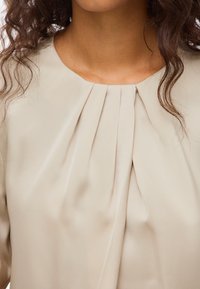 Μπεζ μπλούζα με στρογγυλή λαιμόκοψη, διακριτική πτυχώση κοντά στον γιακά και λεία, μαλακή υφή υφάσματος.