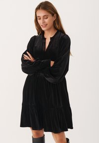 Abito in velluto nero con maniche lunghe e voluminose, scollatura a V e gonna a strati. Il tessuto ha una consistenza liscia con una leggera lucentezza.