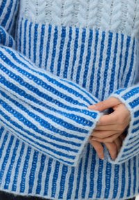 Heleroheline kampsun, tumedamate siniste triipudega. Omaduseks sooniline tekstuur, pikad varrukad ja laiad mansetid karvaste servadega.