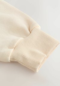 Manche de sweat-shirt crème clair avec poignets côtelés, présentant une texture douce et une finition lisse. Design simple sans éléments métalliques apparents ni motifs.