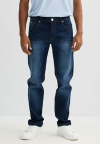 Jeans de mezclilla azul oscuro con corte recto, cinco bolsillos y costuras en contraste. Llevados con zapatillas blancas y una camisa azul clara.