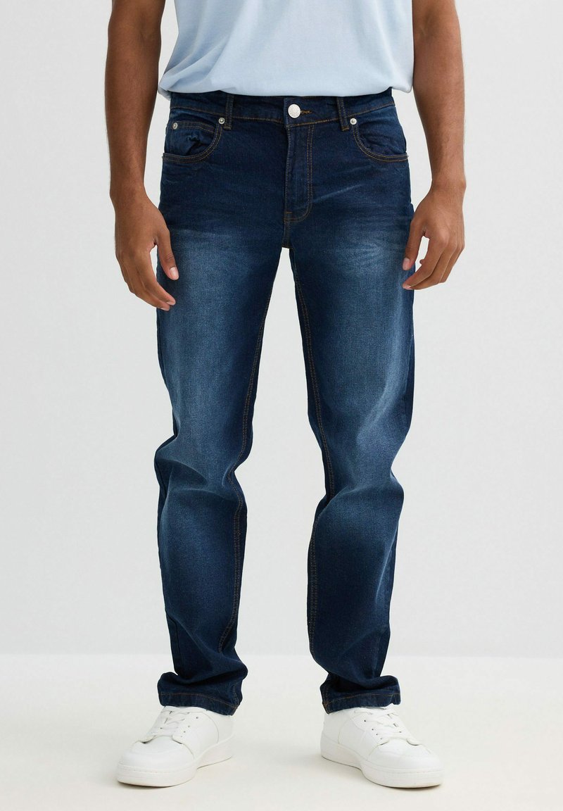 Jeans de mezclilla azul oscuro con corte recto, cinco bolsillos y costuras en contraste. Llevados con zapatillas blancas y una camisa azul clara.