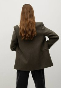 Svetle olivově zelené sako z vlněné směsi se strukturovaným tvarem, šálovým límcem a dvěma postranními kapsami, nošené přes tmavé kalhoty, viděné zezadu.