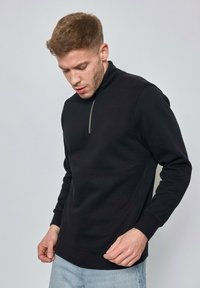 Sweat-shirt à col zippé noir avec manches longues, en tissu doux, au design ajusté et aux poignets côtelés. La fermeture éclair est de couleur argentée contrastante.