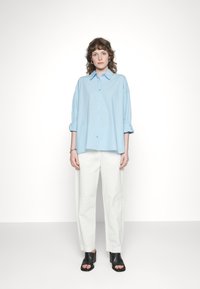 Camisa azul-claro de botões com mangas enroladas, combinada com calças brancas de perna reta e sapatos de salto aberto pretos. Linhas limpas e design minimalista.