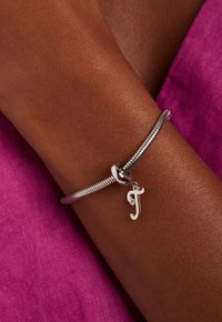 Bracelet en métal argenté avec un design en chaîne de serpent et un petit charm en forme de lettre "j". Le charm présente un accent en diamant. Arrière-plan en tissu rose.