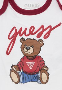 Balts kokvilnas bodijs ar bordo apdari, kas attēlo multfilmu lāci sarkanā džemperī, sēžot ar ziliem džinsiem, un virs tā ir "GUESS" logo.