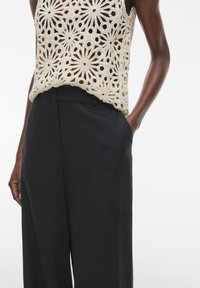 Top de tirantes de ganchillo en beige con patrón floral, combinado con pantalones anchos negros que tienen una textura suave y bolsillos laterales.