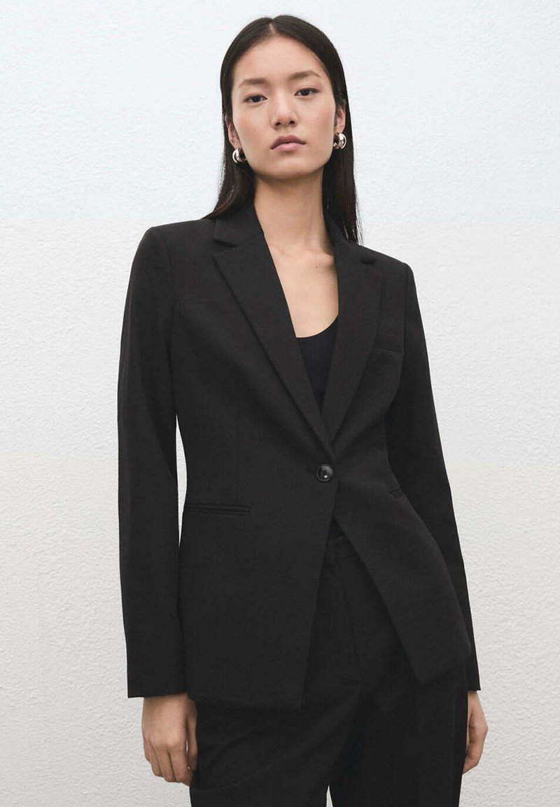 Femme aux longs cheveux raides portant des boucles d'oreilles créoles argentées et une veste de costume noire cintrée sur un haut noir, debout contre un mur clair.