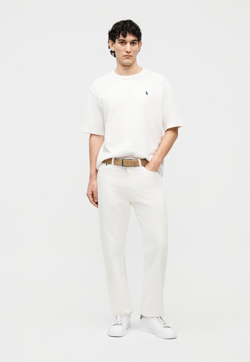 Jonge man staand met een wit T-shirt, witte broek, bruine riem en witte sneakers, tegen een effen witte achtergrond.