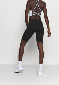 Czarne krótkie spodenki sportowe o dopasowanym kroju, zestawione z białym biustonoszem sportowym z wyraźnym logo na ramiączku. Modelka nosi białe trampki.