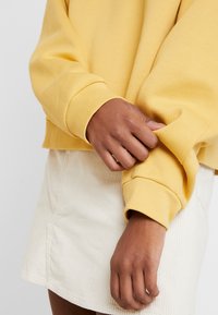 Sudadera amarilla con corte cropped, puños acanalados y textura suave, combinada con una falda de pana de color claro.