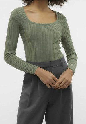 Žena v žebrovaném světle zeleném topu s dlouhým rukávem a hranatým výstřihem, vysokým pasem a tmavě šedých plisovaných kalhotách, ruce sepjaté.